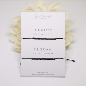 Op de afbeelding: Twee zwarte armbandjes van touw met zwarte kralen die het woord "CUSTOM" in morsecode spellen. De armbandjes worden getoond op een witte kaart met de tekst "Secret message morse code bracelet" en "HANDMADE www.lapalomaboutique.com".