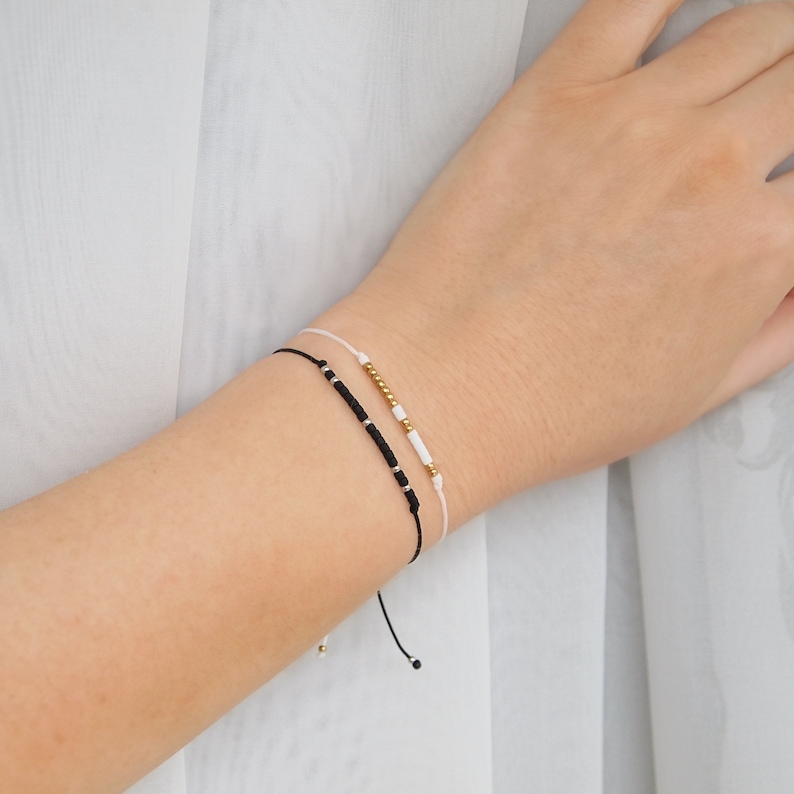 May include: Two friendship bracelets on a wrist. One bracelet is black with gold beads and the other is white with gold beads.