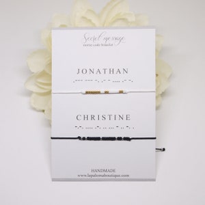 May include: Two white cards with black text. The top card says "Secret message morse code bracelet". The bottom card says "JONATHAN" and "CHRISTINE" with morse code underneath each name. A white bracelet with gold beads is above the top card and a black bracelet with black beads is above the bottom card.
