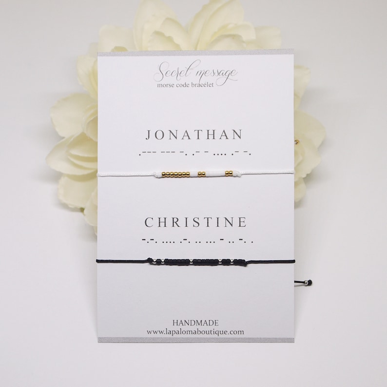 May include: Two white cards with black text. The top card says "Secret message morse code bracelet". The bottom card says "JONATHAN" and "CHRISTINE" with morse code underneath each name. A white bracelet with gold beads is above the top card and a black bracelet with black beads is above the bottom card.