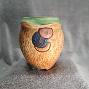 May include: A small, ceramic planter with a textured, orange-brown surface. The planter has a green rim and is decorated with three painted circles in shades of blue, pink, and green.