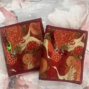 May include: Two rectangular fabric covers featuring a red and orange floral pattern with white crane motifs. Each cover has a dark red fabric border, a metal snap closure, and a fabric loop attached to a small metal charm, one with a green clip.