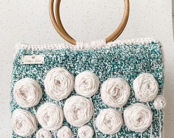 May include: A handmade crochet bag with a wooden handle. The bag is teal and white with white floral embellishments. The bag has a rectangular shape and a light pink trim. The bag has a label that says "Nelly's Knots".