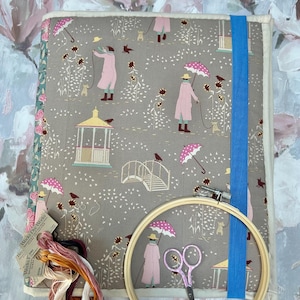 May include: A rectangular fabric project folder with a taupe background featuring a repeating pattern of figures in pink coats with umbrellas, gazebos, and dogs. It includes a blue elastic strap, a wooden embroidery hoop, pink scissors, and embroidery floss.