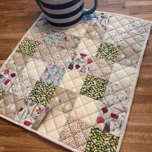 Puede incluir: Un mantel individual acolchado con un diseño de patchwork que presenta varios patrones florales y vegetales en tonos de verde, marrón, blanco y rojo. El mantel individual es beige con un borde blanco y está sobre una superficie de madera. Una tetera a rayas azules y blancas con una tapa rosa y el texto "GOOD LIFE" está encima del mantel individual.