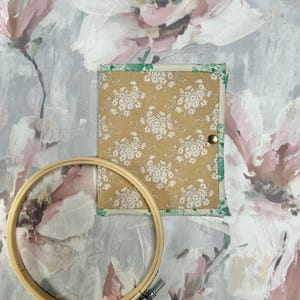 May include: A square fabric case with a mustard yellow background and a white floral pattern, featuring a green floral border and a single metal snap closure, positioned next to a round wooden embroidery hoop with a metal tightening screw.