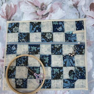 May include: A quilted fabric project bag featuring a blue and cream floral checkerboard pattern, a horizontal zipper closure with a metal dog charm, a wooden embroidery hoop, and small pink-handled scissors resting on the surface.