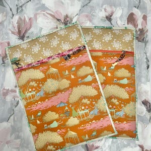May include: Two rectangular fabric pouches featuring a vibrant orange garden scene with trees, gazebos, and figures. Each pouch has a tan floral top panel, a zippered closure with a small metal charm, and a green piped border along the edges.