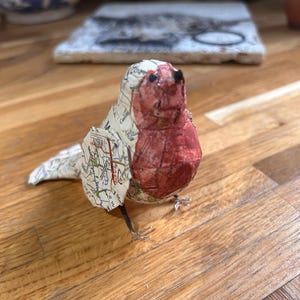 May include: A handmade paper bird sculpture with a red breast and map-printed wings and body. The bird has small metal feet and is positioned on a wooden surface. The sculpture is a unique decorative item.