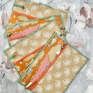 May include: Three rectangular zippered pouches featuring a dual-pattern design. One side displays a tan fabric with a white floral print, while the other side shows an orange landscape scene with figures. Each pouch has a teal floral trim and a metal zipper.
