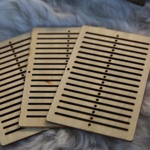 Può includere: Tre telai in legno con fessure per la tessitura. I telai sono in legno marrone chiaro e hanno una finitura naturale.