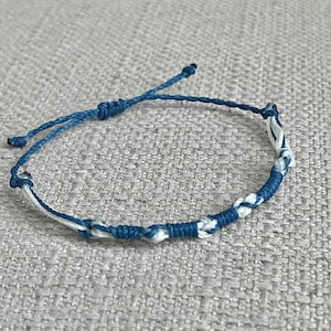 May include: A blue and white braided bracelet. The bracelet is made of woven cord in a pattern of alternating blue and white sections. The ends of the bracelet have adjustable blue cords for sizing. The bracelet is lying on a textured, light gray surface.