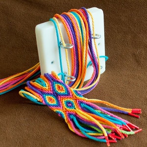 Puede incluir: Un brazalete tejido de colores se está haciendo en un telar de plástico blanco con anillos metálicos. El telar está sujeto a una superficie marrón. El brazalete está hecho con múltiples hilos de lana en tonos de amarillo, naranja, rosa, morado, azul y verde.