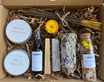 May include: A gift box filled with items for a spiritual cleansing ritual. The box contains white gardenia and palo santo candles, sage smudge spray, a bundle of white sage, a selenite crystal, and a jar of house blessing salts.