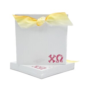 May include: A stack of white notepad paper with a pink "XO" logo. The top notepad has a yellow ribbon tied around it.