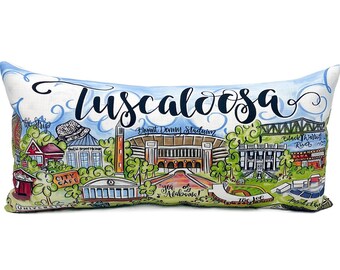 May include: A rectangular pillow with a white background and a colorful illustration of the city of Tuscaloosa, Alabama. The illustration includes landmarks like Bryant Denny Stadium, the Black Warrior River, and the University of Alabama. The text "Tuscaloosa" is written in a large, stylized font at the top of the pillow.