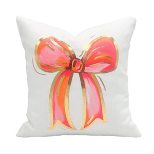 May include: White pillow with a pink and gold bow design. The bow is drawn in a whimsical style with a gold outline.