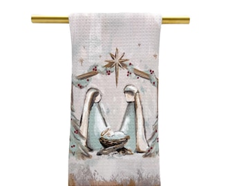 May include: A white kitchen towel with a beige and blue watercolor painting of the nativity scene. The painting features Mary, Joseph, and baby Jesus in a manger with a star above them.