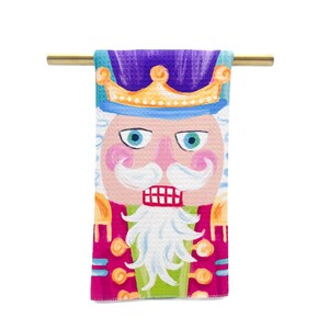 May include: A colorful kitchen towel with a whimsical illustration of a nutcracker. The nutcracker has a purple hat, a gold crown, a pink face, a white beard, and a red mouth. The towel is hanging on a gold rod.