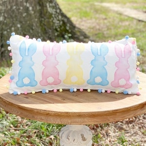 May include: A rectangular white pillow with pastel bunny silhouettes in blue, pink, and yellow. The pillow is trimmed with colorful pom-poms and rests on a round wooden table. The table is supported by a white, ornate pedestal.