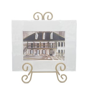 May include: A white framed print of a watercolor painting of a house with a black roof and white columns. The print is on a gold metal easel stand with a decorative scroll design.