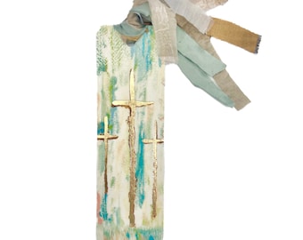 May include: A white bookmark with three gold crosses painted on it. The bookmark has a blue and green watercolor background and is tied with a light blue and beige ribbon.