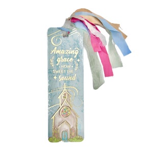 May include: A rectangular bookmark with the text "Amazing grace how sweet the sound" in gold lettering. The bookmark features a church illustration and colorful ribbons in pink, blue, and beige. The background is a light blue color.