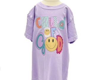 May include: A lilac-colored t-shirt with the words "Child of Good" in a colorful, playful font. The design includes a smiling face and flower accents. The shirt is displayed on a wooden mannequin.