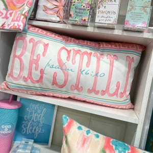 May include: A white pillow with pink lettering that reads "BE STILL Psalm 46:10". The pillow is on a white shelf with other decorative items, including a blue and white patterned throw blanket, a pink and white patterned throw blanket, and a blue and white patterned throw pillow.