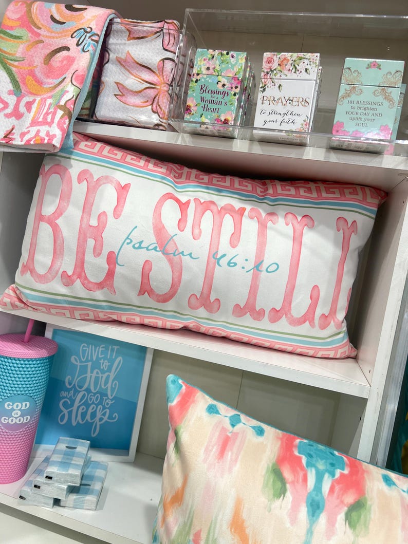 May include: A white pillow with pink lettering that reads "BE STILL Psalm 46:10". The pillow is on a white shelf with other decorative items, including a blue and white patterned throw blanket, a pink and white patterned throw blanket, and a blue and white patterned throw pillow.