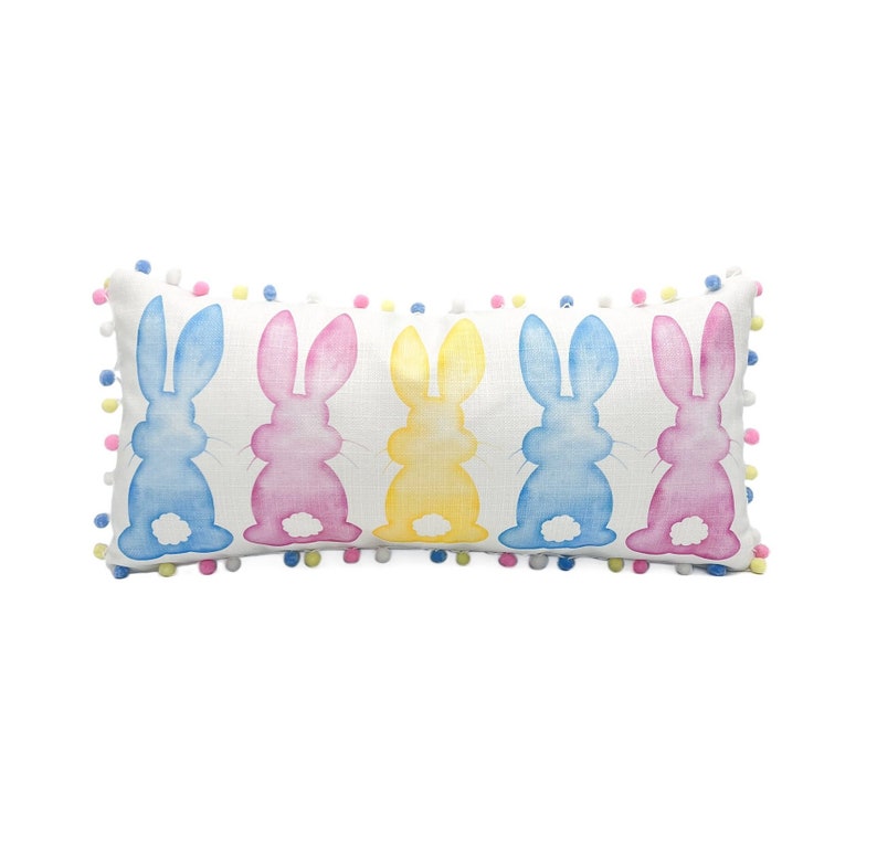 May include: Rectangular decorative pillow featuring five watercolor bunny silhouettes in blue, pink, and yellow. The bunnies are arranged in a row on a white background. The pillow is trimmed with colorful pom-poms, adding a playful touch. Perfect for Easter decor.