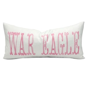 May include: A rectangular white pillow with the words "WAR EAGLE" in pink script across the front. The pillow has a textured appearance and is likely made of linen or a similar fabric. The letters are in a decorative font.