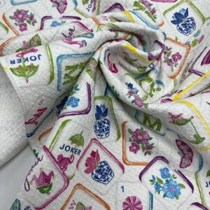 K&ouml;nnte beinhalten: Wei&szlig;er Stoff mit einem farbenfrohen Muster aus Mahjong-Steinen, Blumen und dem Wort "Joker". Die Kacheln sind in verschiedenen Farben umrandet, mit floralen und geometrischen Designs. Der Stoff ist gefaltet und zeigt das sich wiederholende Muster.