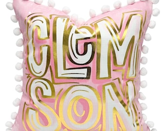 May include: Pink cushion featuring the word "CLEMSON" in gold and white lettering. The cushion has a pink background with white pom-poms around the edges. The letters have a gold outline and a white fill.