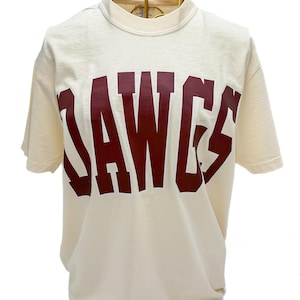 May include: Cream-colored t-shirt with short sleeves and a crew neck. The shirt features the word "DAWGS" in large, maroon block letters across the front. The shirt is displayed on a gold wire mannequin.