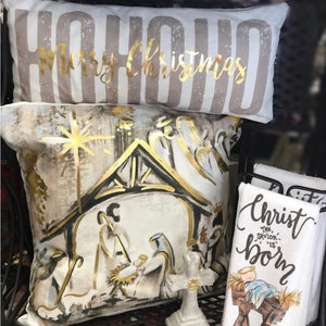 May include: Two decorative throw pillows with a Christmas theme. One pillow is white with gold lettering that says "HOHOHO Merry Christmas". The other pillow is white with a gold and black illustration of the nativity scene.