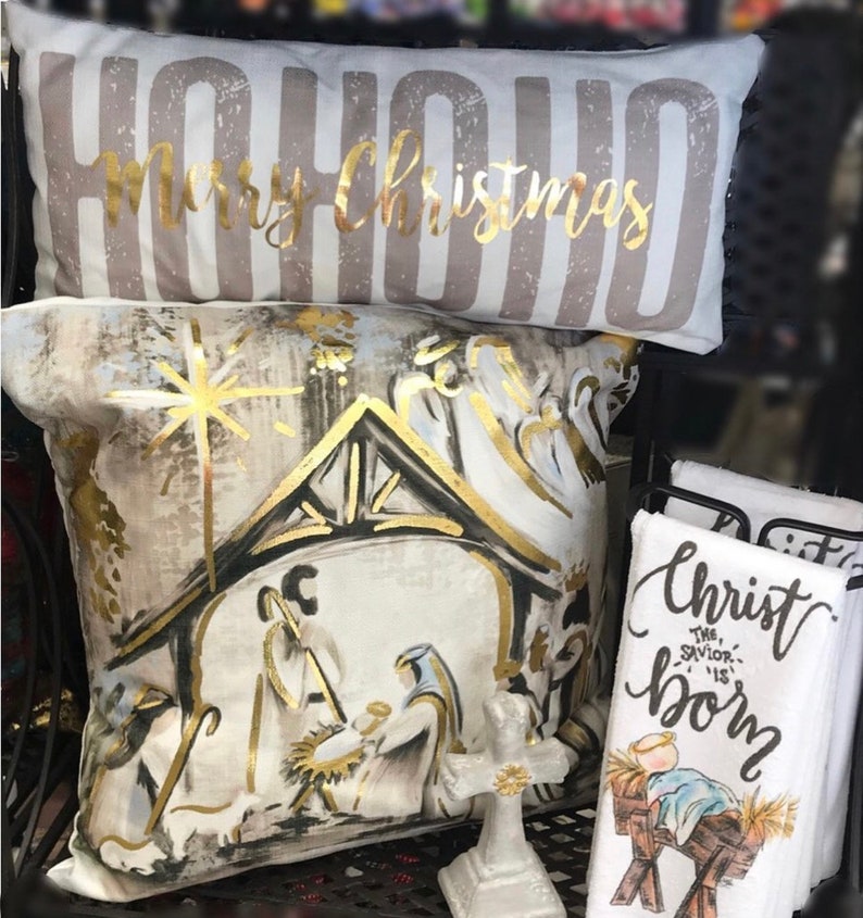 May include: Two decorative throw pillows with a Christmas theme. One pillow is white with gold lettering that says "HOHOHO Merry Christmas". The other pillow is white with a gold and black illustration of the nativity scene.