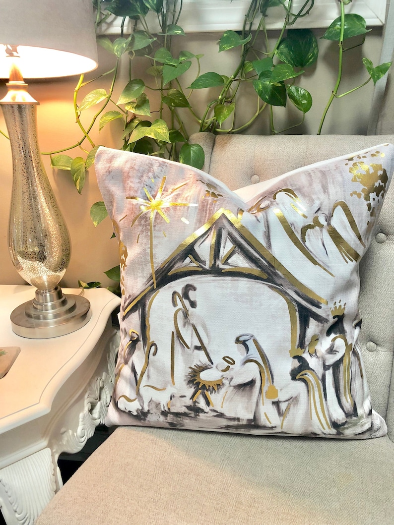 May include: A white throw pillow with a gold and black illustration of the nativity scene. The illustration features a star, a stable, and three figures.