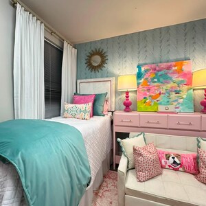 K&ouml;nnte beinhalten: Ein Schlafzimmer mit einem Bett, das eine wei&szlig;e, gesteppte Tagesdecke, eine petrolfarbene Samtdecke und verschiedene rosa und petrolfarbene Kissen hat. Eine rosa Kommode tr&auml;gt zwei rosa Lampen und ein buntes abstraktes Gem&auml;lde. Ein wei&szlig;es Sofa steht davor.