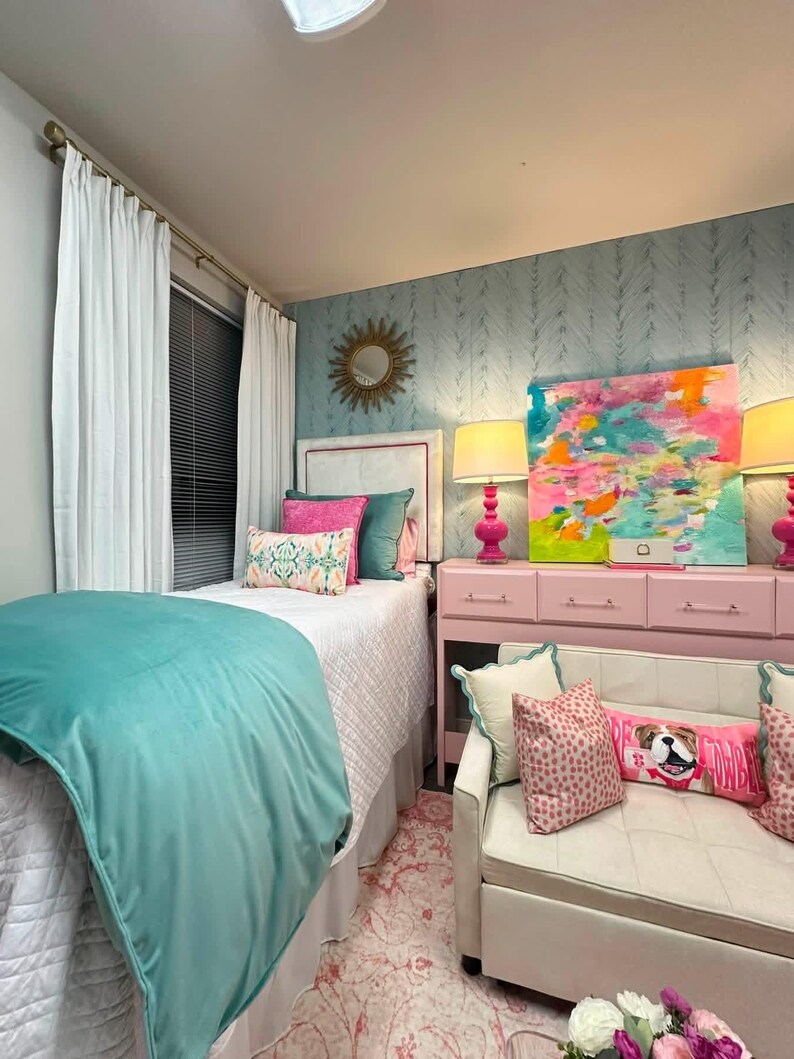 K&ouml;nnte beinhalten: Ein Schlafzimmer mit einem Bett, das eine wei&szlig;e, gesteppte Tagesdecke, eine petrolfarbene Samtdecke und verschiedene rosa und petrolfarbene Kissen hat. Eine rosa Kommode tr&auml;gt zwei rosa Lampen und ein buntes abstraktes Gem&auml;lde. Ein wei&szlig;es Sofa steht davor.