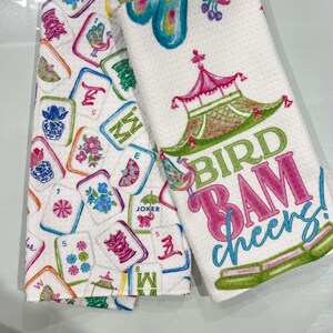K&ouml;nnte beinhalten: Zwei wei&szlig;e K&uuml;chent&uuml;cher. Eines zeigt bunte Mahjong-Kacheln, das andere ein Pagoden-Design mit den Worten "BIRD BAM cheers!". Die Handt&uuml;cher haben eine strukturierte Oberfl&auml;che und sind mit rosa, gr&uuml;nen und blauen Akzenten verziert.