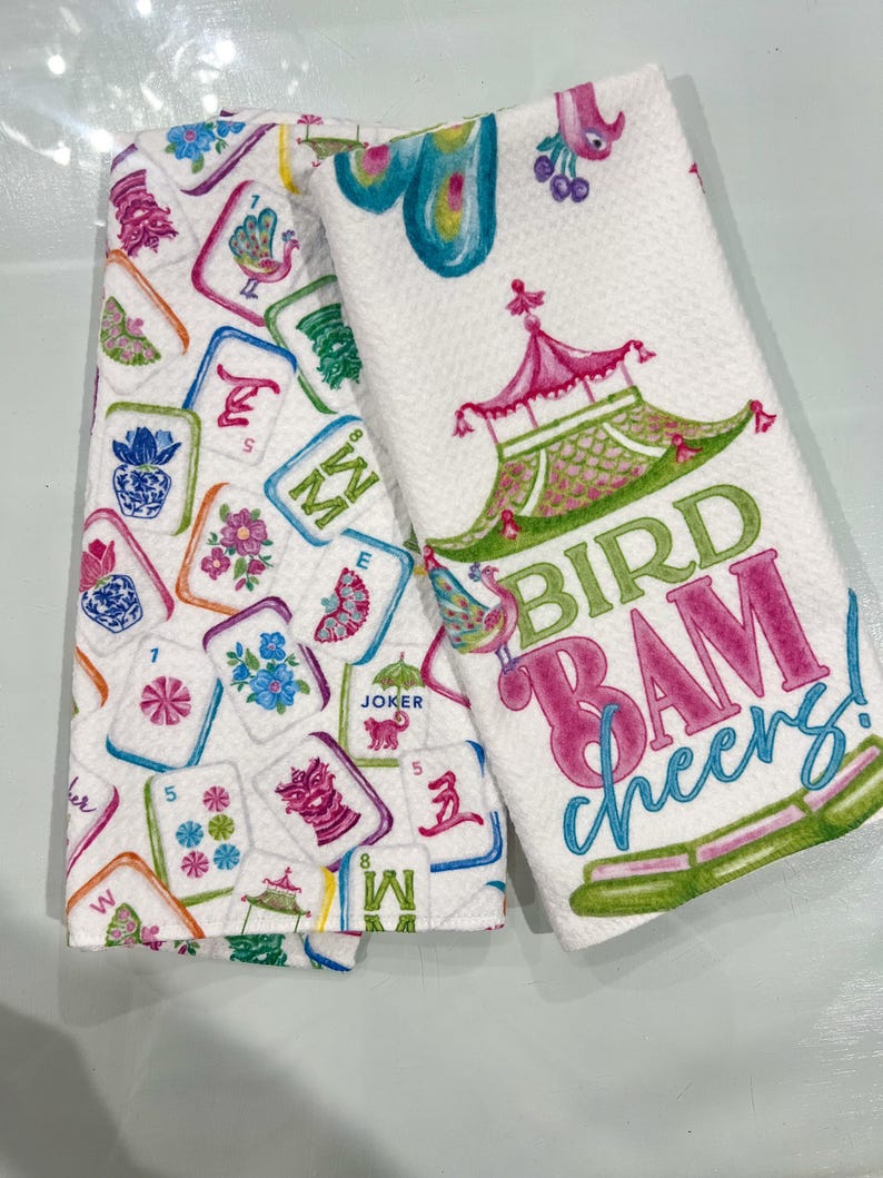 K&ouml;nnte beinhalten: Zwei wei&szlig;e K&uuml;chent&uuml;cher. Eines zeigt bunte Mahjong-Kacheln, das andere ein Pagoden-Design mit den Worten "BIRD BAM cheers!". Die Handt&uuml;cher haben eine strukturierte Oberfl&auml;che und sind mit rosa, gr&uuml;nen und blauen Akzenten verziert.