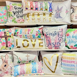 May include: A display of decorative throw pillows with various Easter and spring-themed designs. Pillows feature floral patterns, bunnies, crosses, and phrases like "Home Sweet Home" and "Hoppy Easter." Colors include pastels, gold, and white.