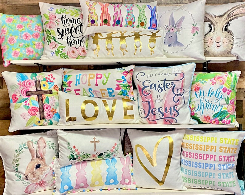 May include: A display of decorative throw pillows with various Easter and spring-themed designs. Pillows feature floral patterns, bunnies, crosses, and phrases like "Home Sweet Home" and "Hoppy Easter." Colors include pastels, gold, and white.