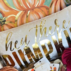 May include: A white velvet pillow with the words "hey there pumpkin" in gold lettering. The pillow is surrounded by fall-themed decorations, including pumpkins and leaves.