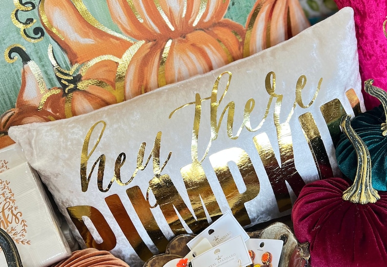 May include: A white velvet pillow with the words "hey there pumpkin" in gold lettering. The pillow is surrounded by fall-themed decorations, including pumpkins and leaves.