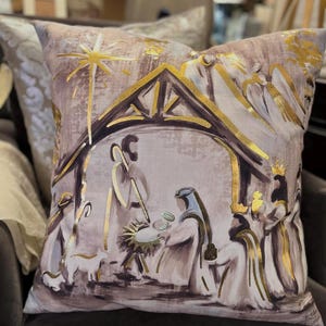 May include: A decorative throw pillow with a beige background and gold accents. The pillow features a painting of the nativity scene with a star above the stable.