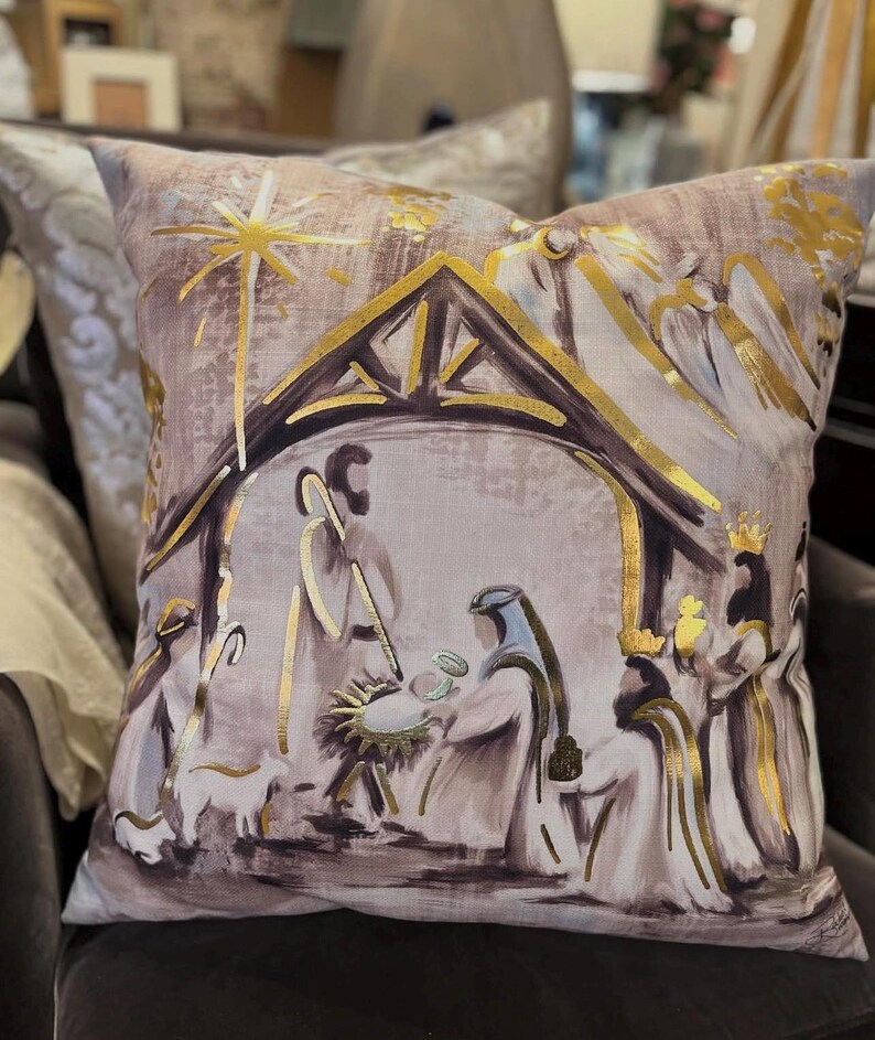 May include: A decorative throw pillow with a beige background and gold accents. The pillow features a painting of the nativity scene with a star above the stable.