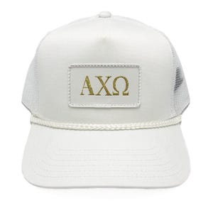 May include: White trucker hat with a mesh back and a curved brim. The hat features a rectangular patch with the gold Greek letters AXΩ. A white rope detail accents the brim.