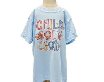 May include: A light blue t-shirt with the words "CHILD OF GOD" in colorful letters and floral accents. The shirt is displayed on a wooden mannequin. The t-shirt has short sleeves and a crew neck.